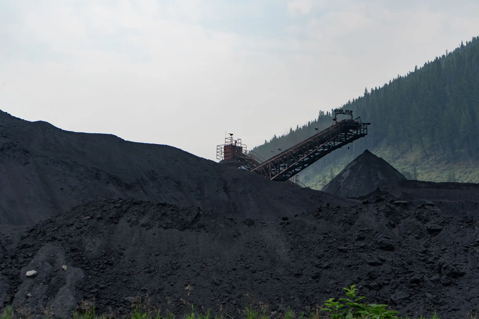 A coal stockpile under a hazy sky, with a stacker conveyor rising over the pile and forested hills behind